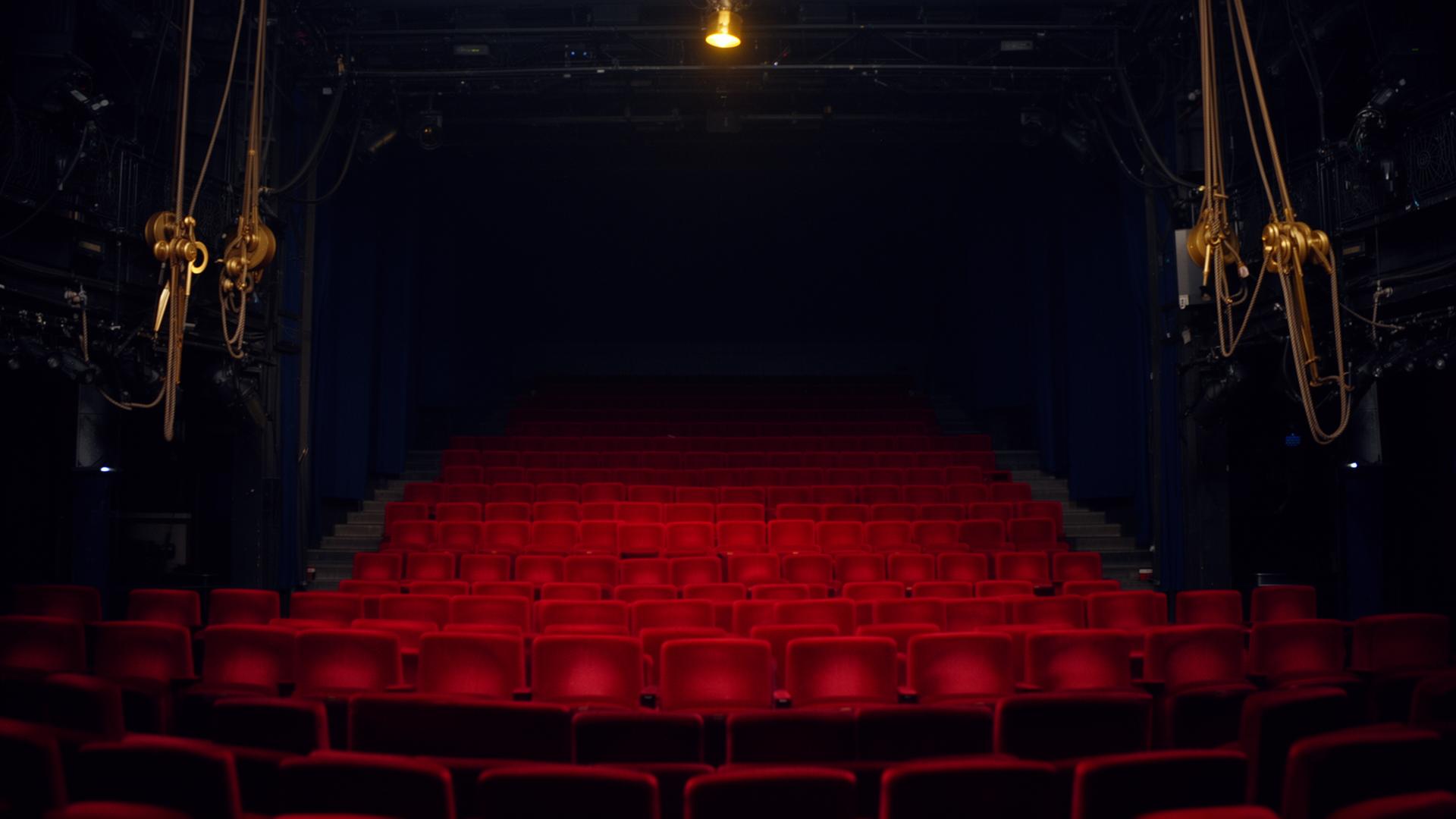 Empty Broadway theatre seen from backstage with a single warm gold stage light and red velvet seats.